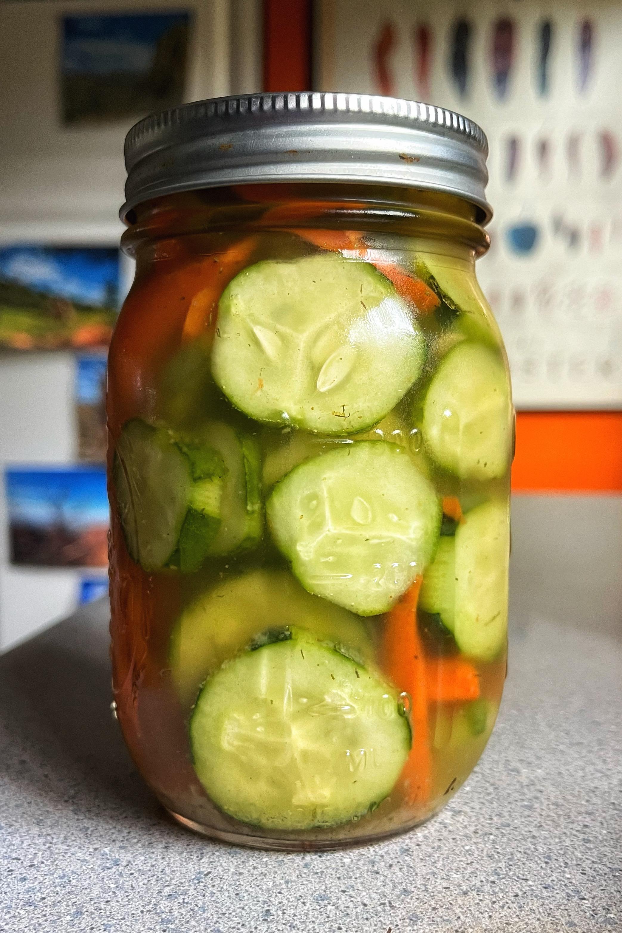 A small glass jar with a metal lid sitting on a gray kitchen countertop. The jar is filled with sliced green cucumbers and orange carrot sticks immersed in a pickling brine.  A small glass jar with a metal lid sitting on a gray kitchen countertop. The jar is filled with sliced green cucumbers and orange carrot sticks immersed in a pickling brine.